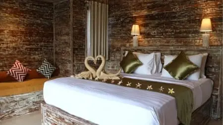 Chambre rustique à Nusa Penida avec grand lit décoré de serviettes en forme de cœur et coussins verts, éclairage mural chaleureux.