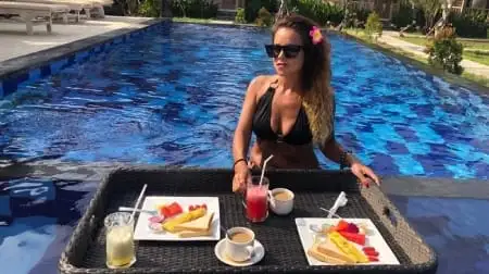 Femme en maillot noir portant des lunettes et une fleur dans les cheveux, assise près d’une piscine à Nusa Penida avec un plateau flottant de petit-déjeuner et boissons.
