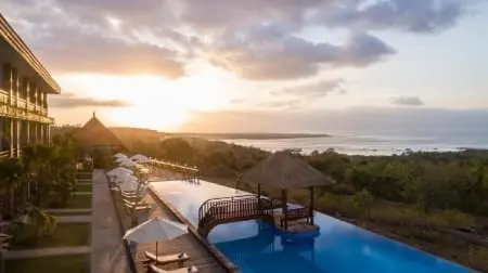 Piscine à débordement au bord d'un hôtel à Nusa Penida au coucher de soleil, avec chaises longues, parasols et petit pont en bois au-dessus de l'eau.