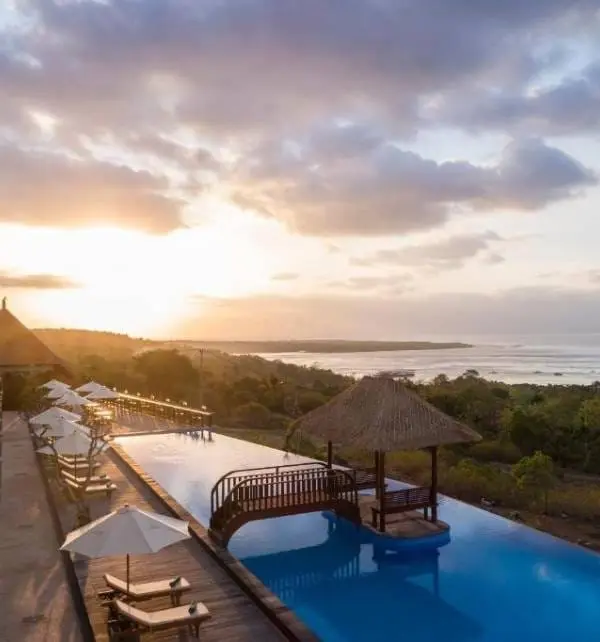 Piscine à débordement au coucher du soleil à Nusa Penida, avec ponton, kiosque en bambou et chaises longues face à la mer.