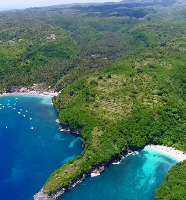 Vue aérienne de Nusa Penida montrant des falaises verdoyantes, des terrasses agricoles et deux criques aux eaux turquoise avec quelques bateaux.