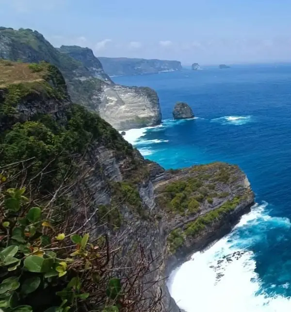 Falaises escarpées de Nusa Penida surplombant l'océan turquoise où les vagues frappent les rochers, vue panoramique côtière en journée.