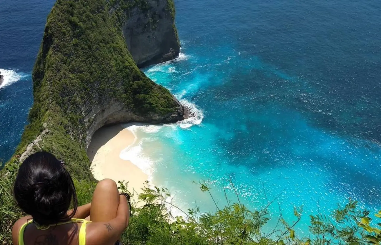Femme assise au sommet d'une falaise regardant la plage en forme de crique et les eaux turquoise de Nusa Penida.