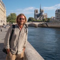 Homme souriant en chemise claire avec sac à dos appuyé sur un parapet au bord de la Seine, cathédrale Notre‑Dame en arrière‑plan.